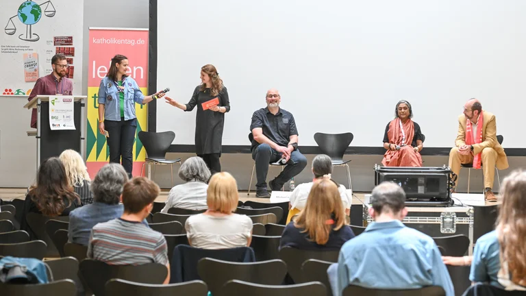 Sechs Personen auf einer B&uuml;hne, drei davon sitzen, die anderen stehen. Eine junge Frau davon &uuml;bergibt einer anderen ein Mikrofon. Im Vordergrund sieht man Menschen im Publikum von hinten.