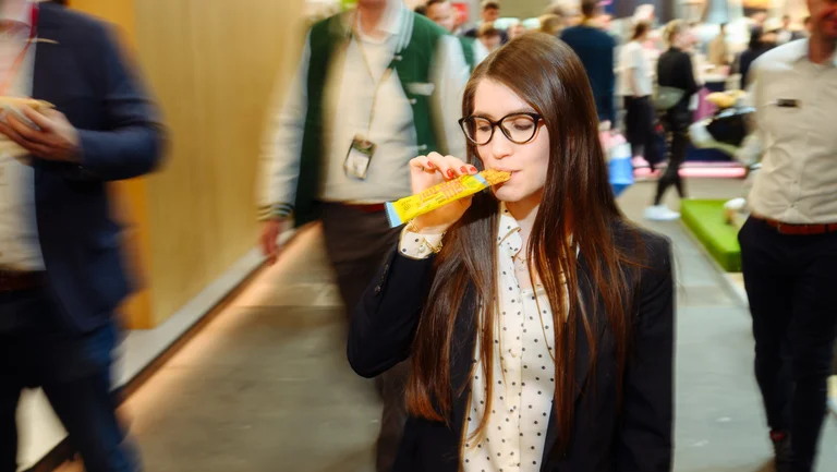 Junge Frau l&auml;uft durch Messehalle und bei&szlig;t vom GEPA-Nussriegel &ldquo;Mango Cocos&rdquo; ab, um sie herum verschwommen weitere Messebesucher.