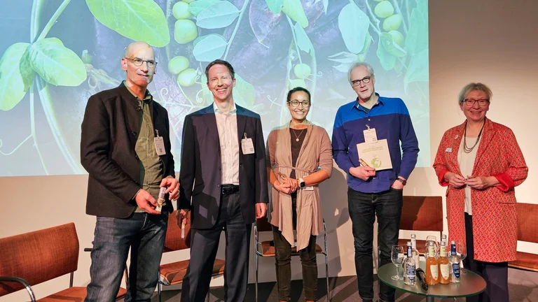 Drei M&auml;nner und zwei Frauen stehen vor einem Stuhlkreis. Hinter ihnen ist ein Bild mit Pflanzen an die Wand projiziert.