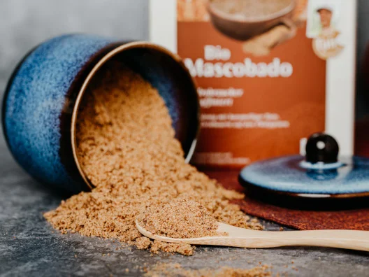 Brauner Zucker l&auml;uft aus einer Zuckerdose auf einen Holzl&ouml;ffel. Im Hintergrund eine Packung GEPA Bio Mascobado