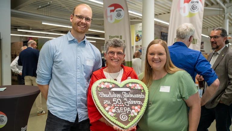 Junger Mann, ältere Frau und junge Frau auf einer Veranstaltung. Die ältere Frau hat ein großes Lebkuchenherz um den Hals mit der Aufschrift „Herzlichen Glückwunsch zu 50. Jahren GEPA“