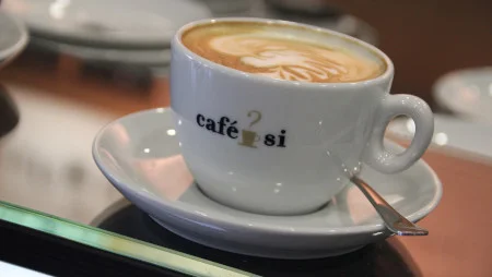 Nahaufnahme einer wei&szlig;e Kaffeetasse mit Kaffee und Latte Art auf einer Untertasse und mit einem L&ouml;ffel. Auf der Tasse steht "Caf&eacute; Si".