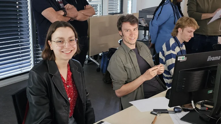 Eine junge Frau und ein junger Mann sitzen an Tischen vor Bildschirmen und lächeln in die Kamera. Um sie herum stehen und sitzen weitere Menschen.