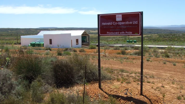 Im Vordergrund steht ein Schild. Darauf steht "Heiveld Co-operative Ltd." und "Produced with Pride". Dahinter ein weißes Gebäude in weiter Landschaft.