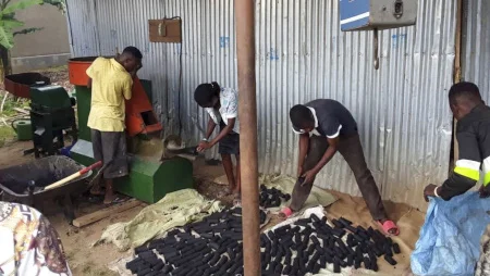 Vier Personen vor einer Wellblechwand stellen mit Hilfe einer Maschine Kohlebriketts her und sammeln sie auf dem Boden vor sich.