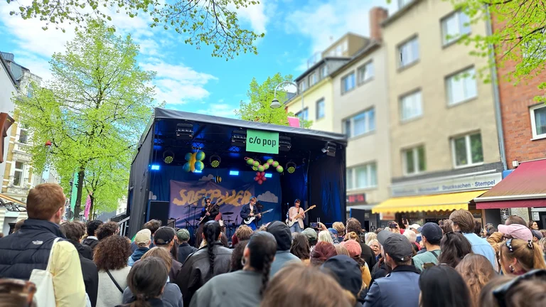 Im Publikum vor einer Open-Air-B&uuml;hne in der Stadt. &Uuml;ber der B&uuml;hne h&auml;ngt ein Schild mit Aufschrift "c/o pop"