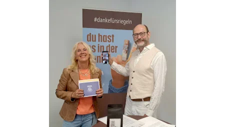 GEPA-Pressesprecherin Barbara Schimmelpfennig und GEPA-Gesch&auml;ftsf&uuml;hrer Peter Schaumberger stehen vor einem GEPA-Banner und l&auml;cheln in die Kamera. Sie h&auml;lt Moderationskarten mit GEPA-Logo, er ein Smartphone.