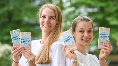 Zwei junge Frauen stehen R&uuml;cken an R&uuml;cken, l&auml;cheln und halten Tafeln der GEPA-Klimaschokolade #Choco4Cahnge in die Kamera.