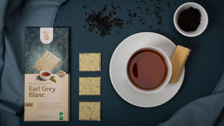 Arrangiertes Bild: Blick von oben auf eine Tafel GEPA-Schokolade "Earl Grey Blanc". Daneben liegen drei ausgepackte St&uuml;cke der Schokolade und eine Schale mit Tee darin.