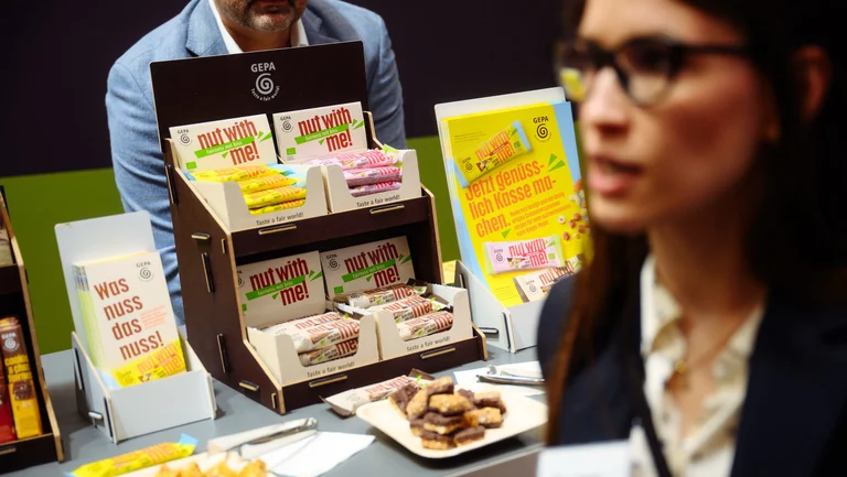Display mit den neuen GEPA-Nussriegeln in allen drei Sorten, im Vordergrund junge Frau im Gespr&auml;ch.