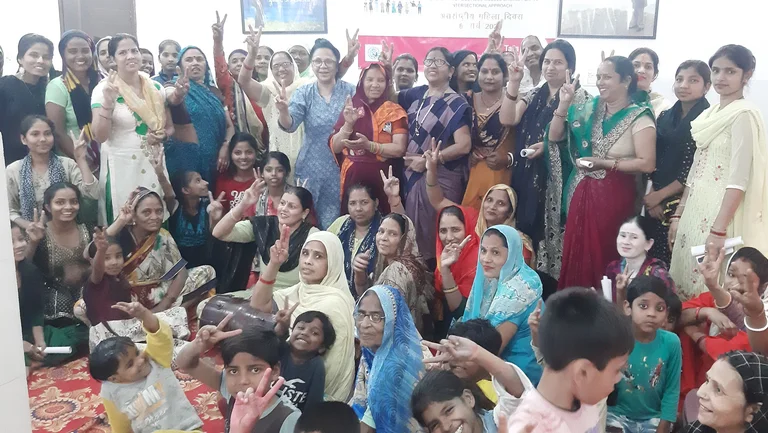 Gruppe von rund 50 Frauen und Kindern, teils mit Saris, l&auml;cheln und gr&uuml;&szlig;en mit Peace-Geste in die Kamera.