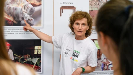 Frau steht vor einer Infotafel mit Bildern und Texten zur Kakaoverarbeitung. Sie deutet auf ein Bild und erkl&auml;rt dabei.