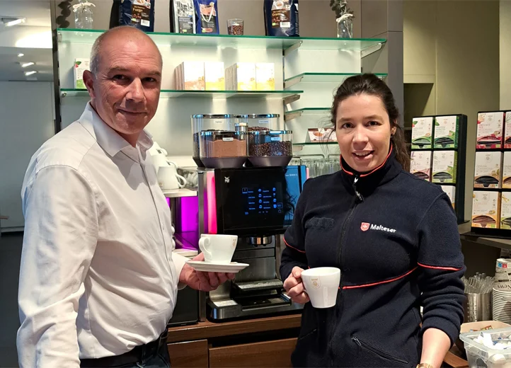 Ein Mann im weißen Hemd und eine Frau im dunklen Malteser-Shirt stehen vor einem Kaffee-Vollautomaten.