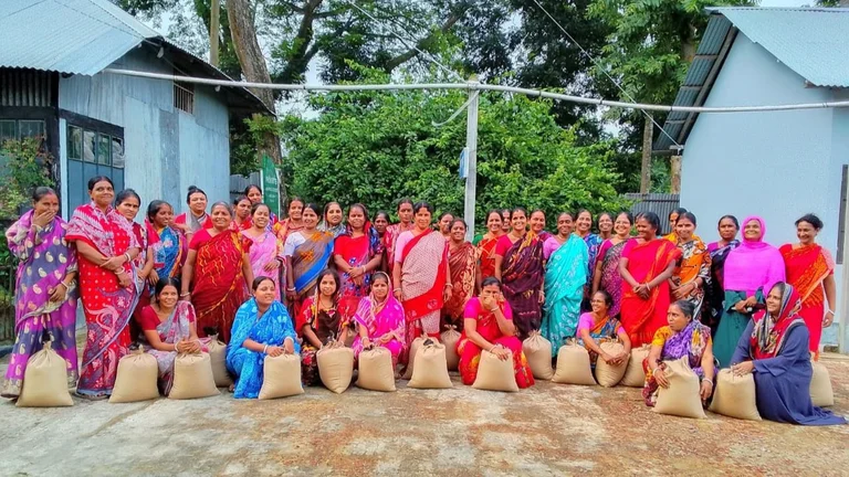 Gruppe von ca. 40 Frauen in Saris l&auml;chelt in die Kamera. Vor ihnen stehen einige S&auml;cke Reis auf dem Boden.