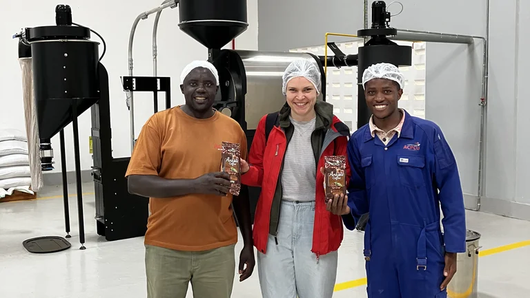 Zwei Männer und eine Frau stehen einem modernen Kaffeeröster und tragen Haarnetze. Sie halten Kaffeepackungen "Ankole Coffee" in die Kamera.