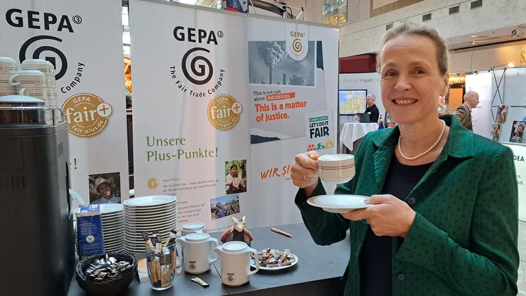 Frau an einem Messestand der GEPA. Sie hält eine Tasse Kaffee und lächelt in die Kamera.
