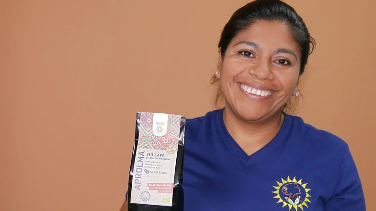Gladyx Hernandez mit blauem APROLMA-Shirt hält lächelnd ein Päckchen in die Kamera.