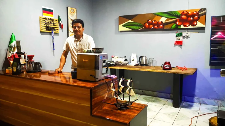 Mann an einer Rezeption. Darauf stehen eine Kaffeemaschine, eine Kanne, eine Packung Kaffee und eine Mexiko-Fahne.