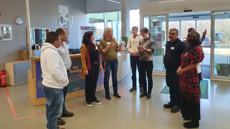 Acht Personen stehen im Halbrund an einem Empfang. Eine Frau erzählt und gestikuliert. Die anderen hören ihr zu.