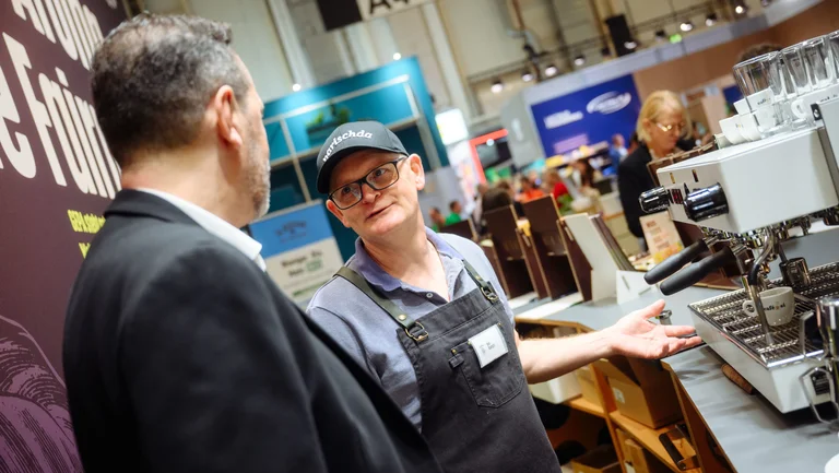 Zwei M&auml;nner unterhalten sich, einer ist Barista und steht hinter einer Siebtr&auml;germaschine.