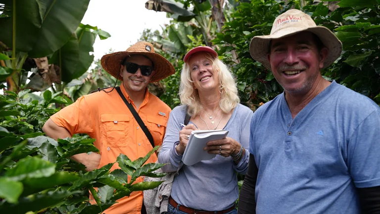 Gruppenbild: zwei M&auml;nner und eine Frau umringt von Kaffeepflanzen lachen in die Kamera. Sie h&auml;lt Stift und Notizblock in der Hand