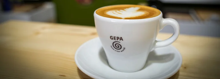 Nahaufnahme einer weißen Kaffeetasse mit GEPA-Logo auf einer Untertasse auf einem Tresen. Auf der Tasse ist Latte Art zu sehen.