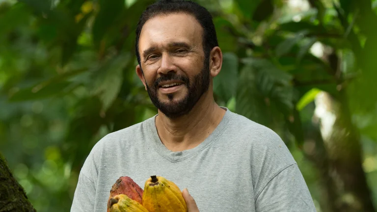Mann h&auml;lt gelbe Kakaobohnen und l&auml;chelt in die Kamera. Hinter ihm ist Gr&uuml;n von B&auml;umen zu erkennen.