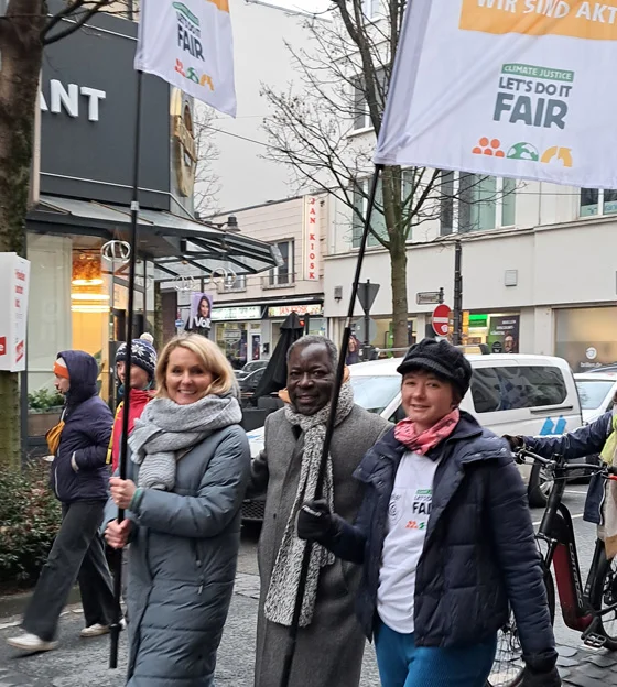3 Personen laufen auf Demo mit, eine trägt eine Fahne mit dem Kampagnenlogo "Climate Justice- Let's Do It Fair"
