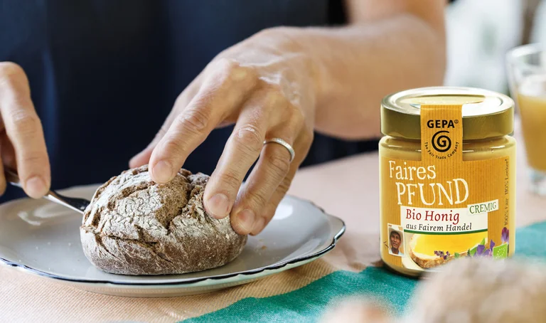 Hände schneiden ein dunkles Brötchen auf, daneben ein Glas GEPA-Honig Faires Pfund