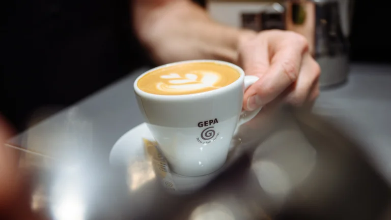 Kaffee mit Latte-Art in wei&szlig;er Tasse mit GEPA-Logo, eine Hand greift nach dem Henkel.