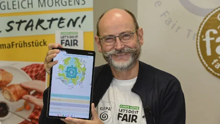 GEPA-Gesch&auml;ftsf&uuml;hrer Peter Schaumberger h&auml;lt ein Tablet ein die Kamera, auf dem das GEPA-Klimapuzzle zu sehen ist. Im Hintergrund sind GEPA-Banner zu sehen.