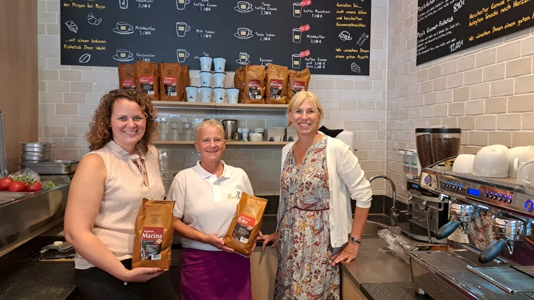 Bild von drei Frauen in der B&auml;ckerei Puppe. Sie l&auml;cheln in die Kamera. Zwei halten je eine Packung GEPA-Kaffee in der Hand.