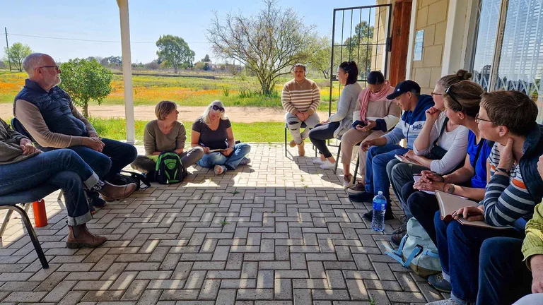 Gruppe von 11 Menschen sitzt auf einer Veranda im Kreis, teils auf Stühlen teils auf dem Boden. Manche notieren etwas in Notizbüchern.
