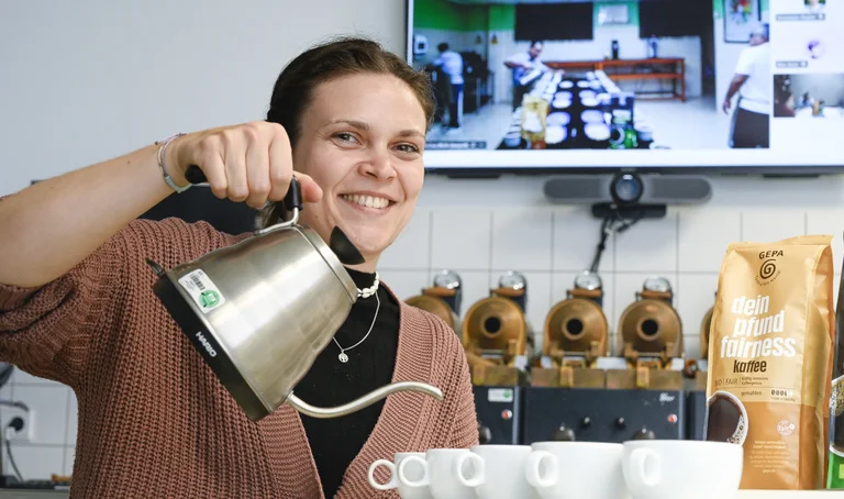 Junge Frau in einem Kaffeelabor hält eine silberne Kanne über weiße Tassen, als würde sie Kaffee aufgießen, und lächelt in die Kamera. Am Rand steht eine Packung GEPA-Kaffee "dein pfund fairness".