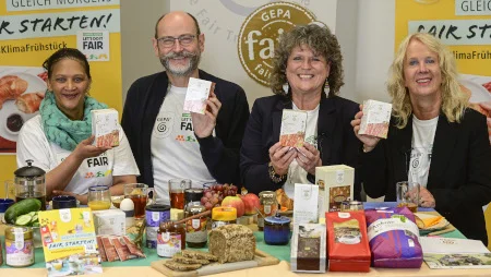Drei Frauen und ein Mann an einem &uuml;ppig bef&uuml;llten Fr&uuml;hst&uuml;ckstisch inkl. GEPA-Produkte lachen und halten GEPA-Teepackungen in die Kamera. Hinter ihnen sind GEPA-Banner aufgestellt.