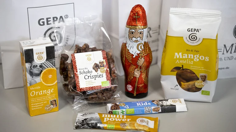 Vor weißen Geschenktüten mit GEPA-Logo stehen GEPA-Produkte: Orangensaft, Schokonikolaus, Mangos, Schoko Crispies, ein Schokoriegel und ein Fruchtriegel.