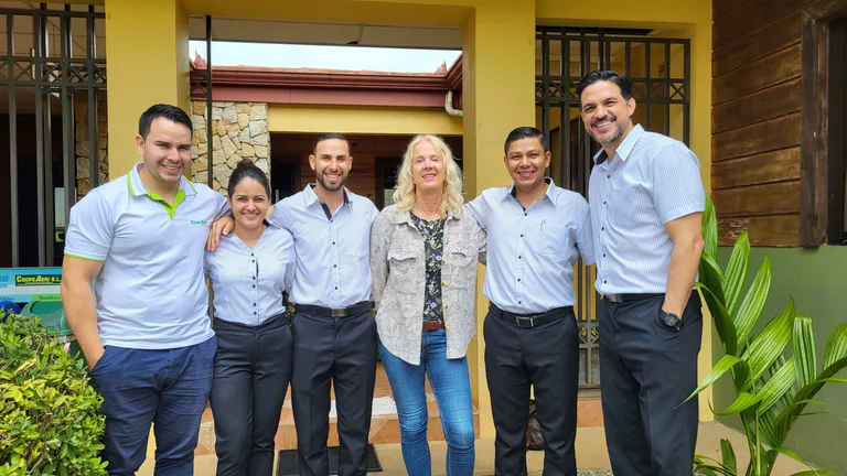 Gruppenbild: vier M&auml;nner und zwei Frauen legen die Arme &uuml;ber die Schultern des Nachbarn. Sie stehen vor dem Eingang eines Geb&auml;udes und l&auml;cheln in die Kamera.