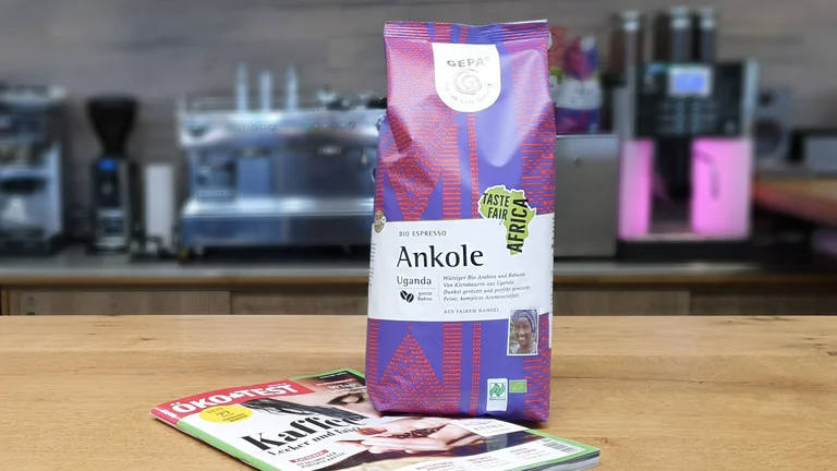 Auf einer Theke steht eine Packung GEPA-Kaffee Bio Espresso Ankole auf einer Zeitschrift ÖKO-TEST. Im Hintergrund sieht man verschwommen Kaffeemaschinen und Automaten.