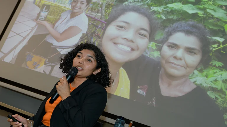 Junge Frau spricht in ein Mikrofon. Auf einer Leinwand hinter ihr ist sie selbst mit einer &auml;lteren Frau zu sehen.