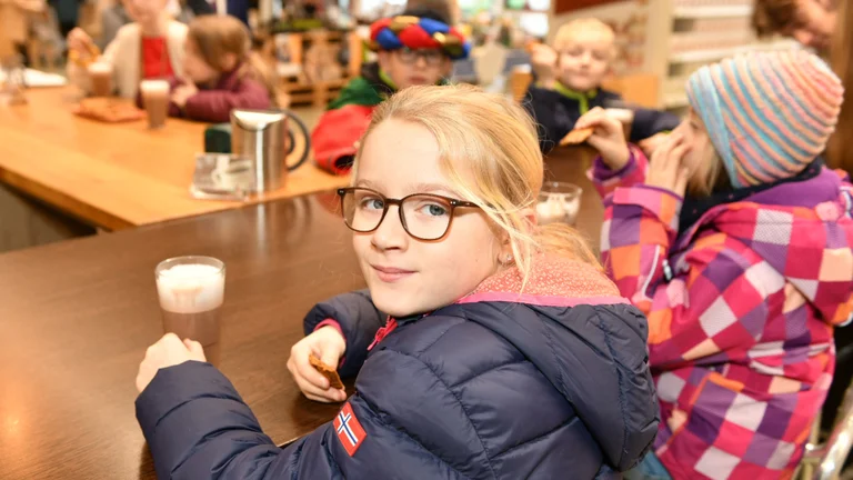 Mädchen in Jacke sitzt an einem Tisch und dreht sich in die Kamera. Vor ihr ein Glas mit Heißer Schokolade. Im Hintergrund sieht man verschwommen weitere Kinder am selben Tisch sitzen.