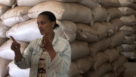 Frau in einer Lagerhalle vor stapelweise S&auml;cken. Sie spricht und gestikuliert zu jemandem abseits der Kamera.