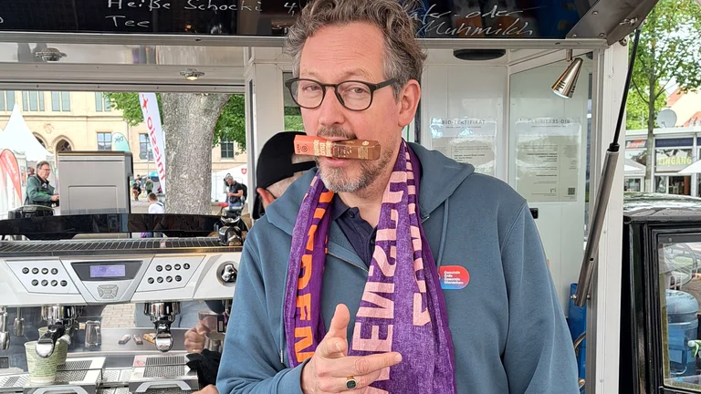 Eckart von Hirschhausen vor einem Kaffeestand im Freien. Er zeigt in die Kamera und hat dabei einen noch verpackten GEPA-Schokoriegel im Mund.
