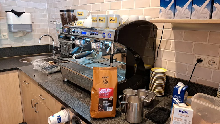 Siebtr&auml;germaschine in einer B&auml;ckereik&uuml;che. Darauf stehen Tassen mit Logo der B&auml;ckerei Puppe. An die Maschine gelehnt steht eine Packung GEPA-Kaffee "Rico"