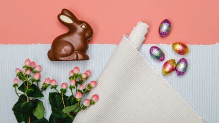 Sicht von oben auf einen Schoko-Osterhasen auf einem Tischset. Daneben liegen dekorativ Blumen und bunte Schokoladen-Ostereier.