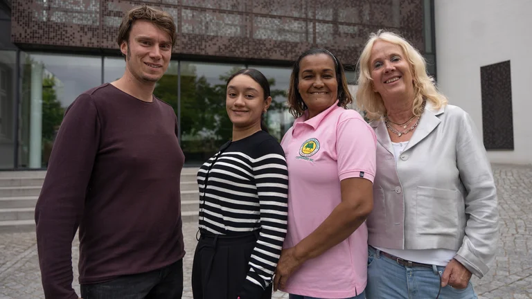 Ein Mann und drei Frauen posieren vor einem Geb&auml;ude und l&auml;cheln in die Kamera.