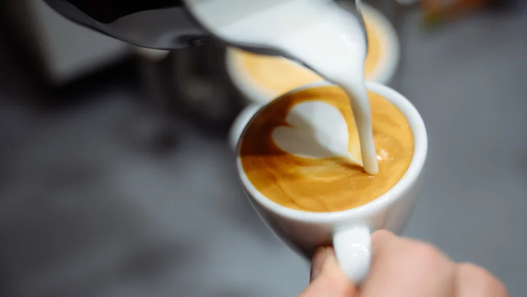 Milch flie&szlig;t aus einem K&auml;nnchen in eine Kaffeetasse, sodass sich ein Herz darauf bildet.