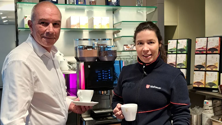 Ein Mann im weißen Hemd und eine Frau im dunklen Malteser-Shirt stehen vor einem Kaffee-Vollautomaten.