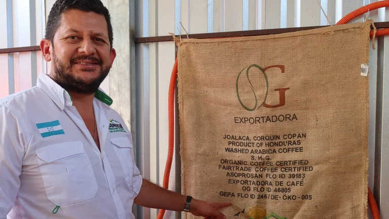 Mann deutet auf einen Kaffeesack mit spanischer Aufschrift, der an einer Wand hängt. Auf seinem weißen Hemd ist die Flagge von Honduras.