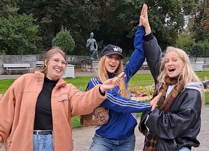 Drei junge Frauen in einem Park. Eine hält den anderen beiden ein Mikro hin. Die beiden machen gemeinsam eine Yogapose. Alle lächeln in die Kamera.