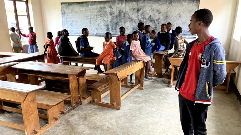 Blick in einen Klassenraum der Nyandozo Primary School in Uganda. Kinder sitzen auf Tischen und Bänken vor einer Tafel.
